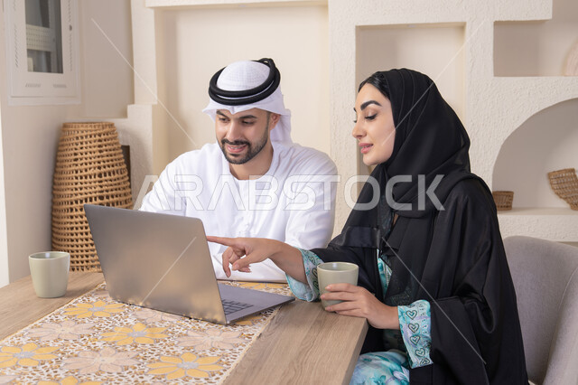 الاشارة الى اللابتوب، العمل عن بعد، إنجاز المهام من المنزل، دمج التقنية والتكنولوجيا في الروتين اليومي، زوجان عربيان خليجيان اماراتيان يجلسان في البيت يعملان على الحاسب المحمول، ايماءات وتعابير التركيز والسرور، تناول المشروبات الساخنة، الترابط والتواصل الاسري