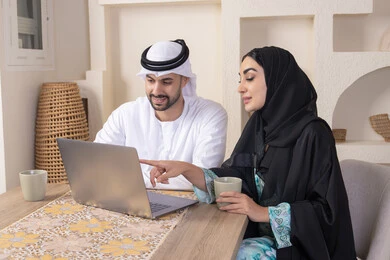 Pointing to laptop, working remotely, completing tasks from home, incorporating technology into daily routine, an Arab Gulf Emirati couple sitting at home working on a laptop, gestures and expressions of concentration and pleasure, drinking hot drinks, family bonding and communication