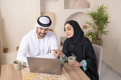 Pointing to laptop, working remotely, completing tasks from home, incorporating technology into daily routine, an Arab Gulf Emirati couple sitting at home working on a laptop, gestures and expressions of concentration and pleasure, drinking hot drinks, family bonding and communication