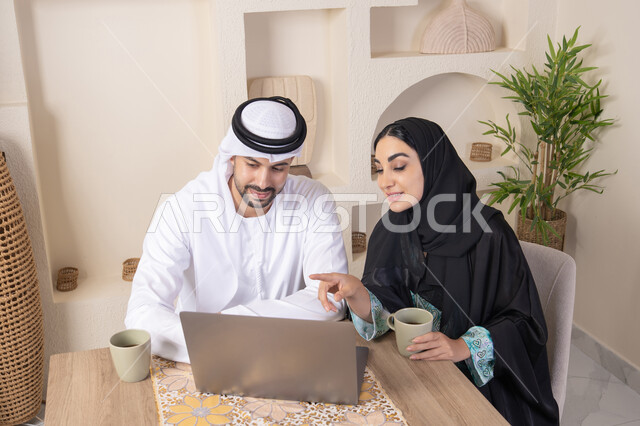 إنجاز المهام من المنزل، دمج التقنية والتكنولوجيا في الروتين اليومي، زوجان عربيان خليجيان اماراتيان يجلسان في البيت يعملان على الحاسب المحمول، ايماءات وتعابير التركيز والسرور، تناول المشروبات الساخنة، الترابط والتواصل الاسري، الاشارة الى اللابتوب، العمل عن بعد