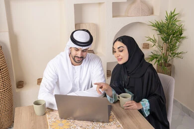 Getting things done from home, incorporating technology into daily routine, an Emirati Gulf Arab couple sitting at home working on a laptop, gestures and expressions of focus and pleasure, drinking hot drinks, family bonding and communication, pointing at the laptop, working remotely