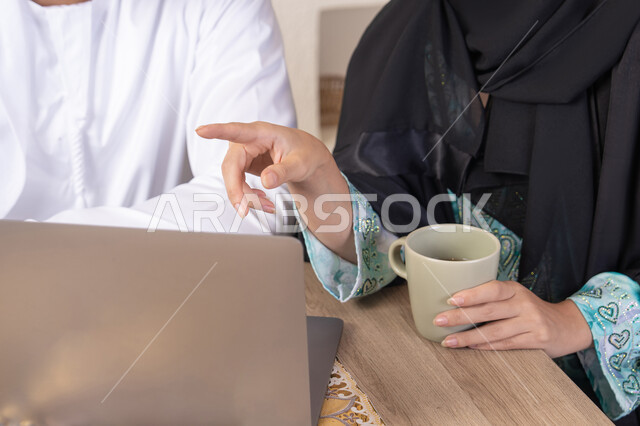 Pointing to laptop, working remotely, completing tasks from home, incorporating technology into daily routine, an Arab Gulf Emirati couple sitting at home working on a laptop, gestures and expressions of concentration and pleasure, drinking hot drinks, family bonding and communication