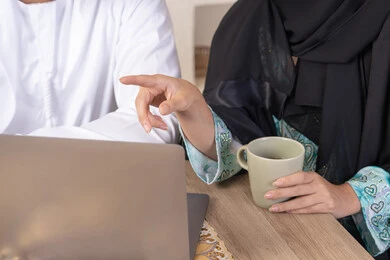 الاشارة الى اللابتوب، العمل عن بعد، إنجاز المهام من المنزل، دمج التقنية والتكنولوجيا في الروتين اليومي، زوجان عربيان خليجيان اماراتيان يجلسان في البيت يعملان على الحاسب المحمول، ايماءات وتعابير التركيز والسرور، تناول المشروبات الساخنة، الترابط والتواصل الاسري