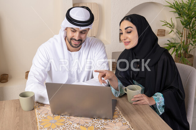 إنجاز المهام من المنزل، دمج التقنية والتكنولوجيا في الروتين اليومي، زوجان عربيان خليجيان اماراتيان يجلسان في البيت يعملان على الحاسب المحمول، ايماءات وتعابير التركيز والسرور، تناول المشروبات الساخنة، الترابط والتواصل الاسري، الاشارة الى اللابتوب، العمل عن بعد
