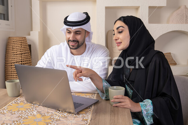 الاشارة الى اللابتوب، العمل عن بعد، إنجاز المهام من المنزل، دمج التقنية والتكنولوجيا في الروتين اليومي، زوجان عربيان خليجيان اماراتيان يجلسان في البيت يعملان على الحاسب المحمول، ايماءات وتعابير التركيز والسرور، تناول المشروبات الساخنة، الترابط والتواصل الاسري