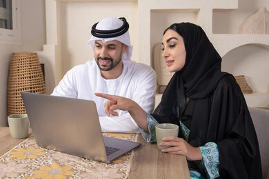الاشارة الى اللابتوب، العمل عن بعد، إنجاز المهام من المنزل، دمج التقنية والتكنولوجيا في الروتين اليومي، زوجان عربيان خليجيان اماراتيان يجلسان في البيت يعملان على الحاسب المحمول، ايماءات وتعابير التركيز والسرور، تناول المشروبات الساخنة، الترابط والتواصل الاسري