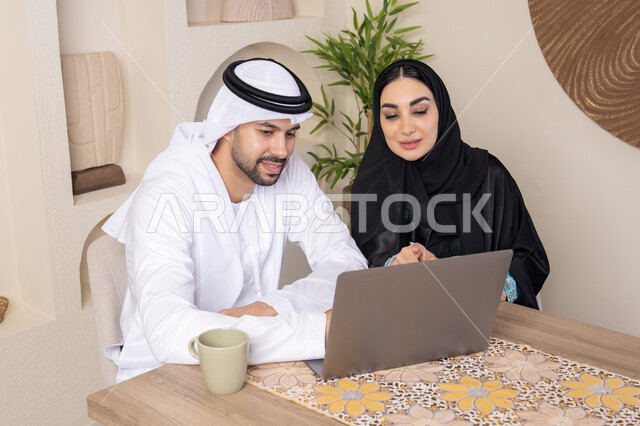 إنجاز المهام من المنزل، دمج التقنية والتكنولوجيا في الروتين اليومي، زوجان عربيان خليجيان اماراتيان يجلسان في البيت يعملان على الحاسب المحمول، ايماءات وتعابير التركيز والسرور، تناول المشروبات الساخنة، الترابط والتواصل الاسري، الاشارة الى اللابتوب، العمل عن بعد
