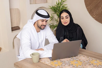 Getting things done from home, incorporating technology into daily routine, an Emirati Gulf Arab couple sitting at home working on a laptop, gestures and expressions of focus and pleasure, drinking hot drinks, family bonding and communication, pointing at the laptop, working remotely