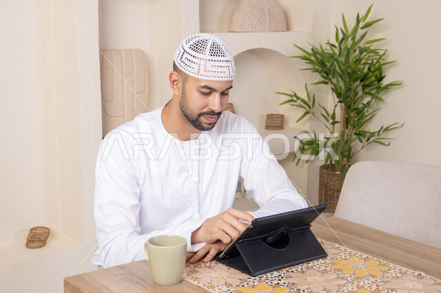 استخدام جهاز تقني حديث متطور، الاستمتاع بتناول المشروبات الساخنة، أداء المهام من المنزل، العمل عن بعد، رجل عربي خليجي اماراتي يرتدي الكندورة والطاقية البيضاء يجلس على الطاولة يعمل على جهاز التابلت، دمج التقنية والتكنولوجيا في الحياة اليومية