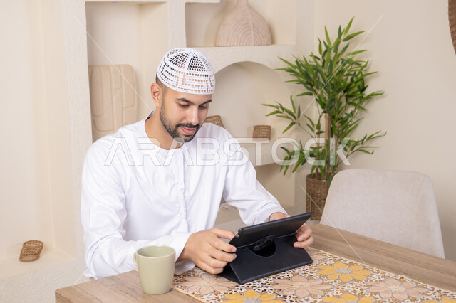 أداء المهام من المنزل، العمل عن بعد، رجل عربي خليجي اماراتي يرتدي الكندورة والطاقية البيضاء يجلس على الطاولة يعمل على جهاز التابلت، دمج التقنية والتكنولوجيا في الحياة اليومية، استخدام جهاز تقني حديث متطور، الاستمتاع بتناول المشروبات الساخنة