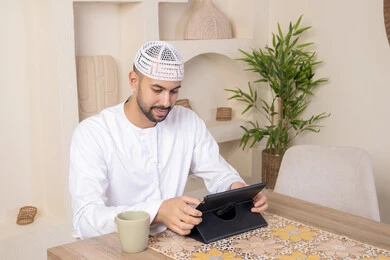 أداء المهام من المنزل، العمل عن بعد، رجل عربي خليجي اماراتي يرتدي الكندورة والطاقية البيضاء يجلس على الطاولة يعمل على جهاز التابلت، دمج التقنية والتكنولوجيا في الحياة اليومية، استخدام جهاز تقني حديث متطور، الاستمتاع بتناول المشروبات الساخنة