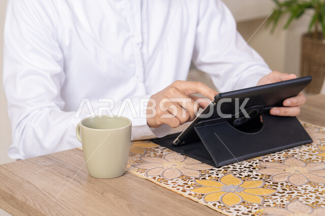 استخدام جهاز تقني حديث متطور، الاستمتاع بتناول المشروبات الساخنة، أداء المهام من المنزل، العمل عن بعد، رجل عربي خليجي اماراتي يرتدي الكندورة والطاقية البيضاء يجلس على الطاولة يعمل على جهاز التابلت، دمج التقنية والتكنولوجيا في الحياة اليومية