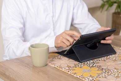 استخدام جهاز تقني حديث متطور، الاستمتاع بتناول المشروبات الساخنة، أداء المهام من المنزل، العمل عن بعد، رجل عربي خليجي اماراتي يرتدي الكندورة والطاقية البيضاء يجلس على الطاولة يعمل على جهاز التابلت، دمج التقنية والتكنولوجيا في الحياة اليومية
