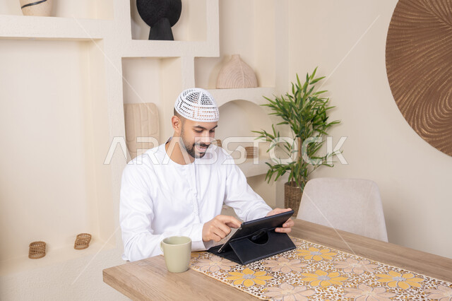 إتمام المهام من المنزل، العمل عن بعد، رجل عربي خليجي اماراتي يرتدي الكندورة والطاقية البيضاء يجلس على الطاولة يعمل على جهاز التابلت، دمج التقنية والتكنولوجيا في الحياة اليومية، استخدام جهاز تقني حديث متطور، الاستمتاع بتناول المشروبات الساخنة