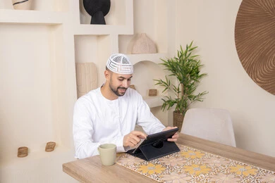 إتمام المهام من المنزل، العمل عن بعد، رجل عربي خليجي اماراتي يرتدي الكندورة والطاقية البيضاء يجلس على الطاولة يعمل على جهاز التابلت، دمج التقنية والتكنولوجيا في الحياة اليومية، استخدام جهاز تقني حديث متطور، الاستمتاع بتناول المشروبات الساخنة