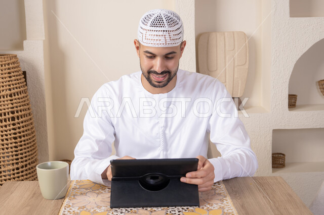 أداء المهام من المنزل، العمل عن بعد، رجل عربي خليجي اماراتي يرتدي الكندورة والطاقية البيضاء يجلس على الطاولة يعمل على جهاز التابلت، دمج التقنية والتكنولوجيا في الحياة اليومية، استخدام جهاز تقني حديث متطور، الاستمتاع بتناول المشروبات الساخنة