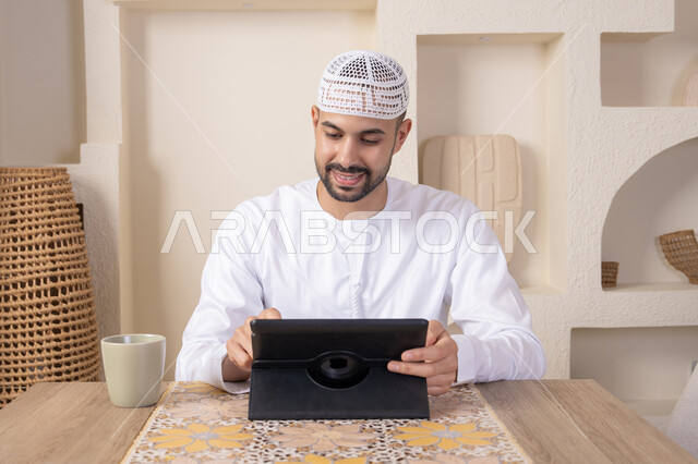 استخدام جهاز تقني حديث متطور، الاستمتاع بتناول المشروبات الساخنة، أداء المهام من المنزل، العمل عن بعد، رجل عربي خليجي اماراتي يرتدي الكندورة والطاقية البيضاء يجلس على الطاولة يعمل على جهاز التابلت، دمج التقنية والتكنولوجيا في الحياة اليومية
