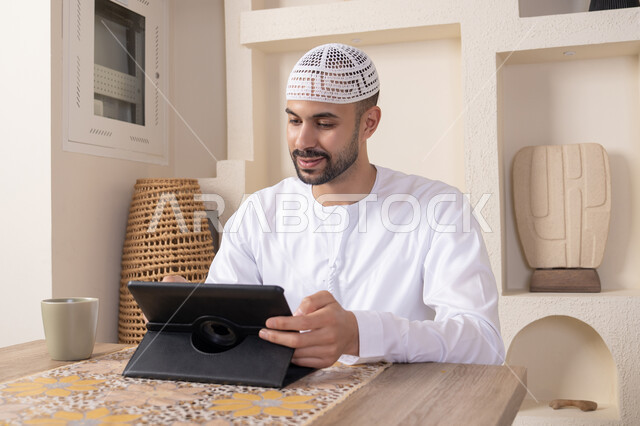 إتمام المهام من المنزل، العمل عن بعد، رجل عربي خليجي اماراتي يرتدي الكندورة والطاقية البيضاء يجلس على الطاولة يعمل على جهاز التابلت، دمج التقنية والتكنولوجيا في الحياة اليومية، استخدام جهاز تقني حديث متطور، الاستمتاع بتناول المشروبات الساخنة