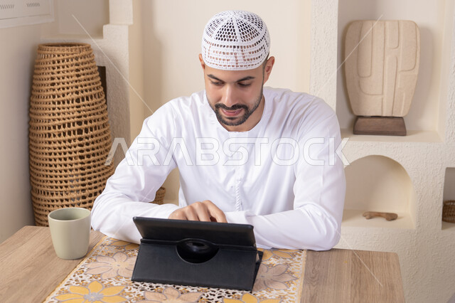أداء المهام من المنزل، العمل عن بعد، رجل عربي خليجي اماراتي يرتدي الكندورة والطاقية البيضاء يجلس على الطاولة يعمل على جهاز التابلت، دمج التقنية والتكنولوجيا في الحياة اليومية، استخدام جهاز تقني حديث متطور، الاستمتاع بتناول المشروبات الساخنة