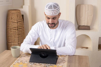 أداء المهام من المنزل، العمل عن بعد، رجل عربي خليجي اماراتي يرتدي الكندورة والطاقية البيضاء يجلس على الطاولة يعمل على جهاز التابلت، دمج التقنية والتكنولوجيا في الحياة اليومية، استخدام جهاز تقني حديث متطور، الاستمتاع بتناول المشروبات الساخنة