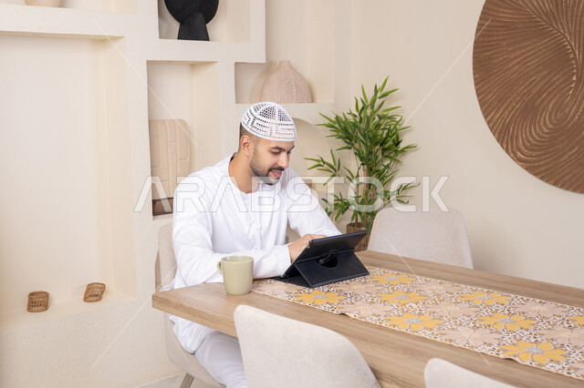 استخدام جهاز تقني حديث متطور، الاستمتاع بتناول المشروبات الساخنة، أداء المهام من المنزل، العمل عن بعد، رجل عربي خليجي اماراتي يرتدي الكندورة والطاقية البيضاء يجلس على الطاولة يعمل على جهاز التابلت، دمج التقنية والتكنولوجيا في الحياة اليومية