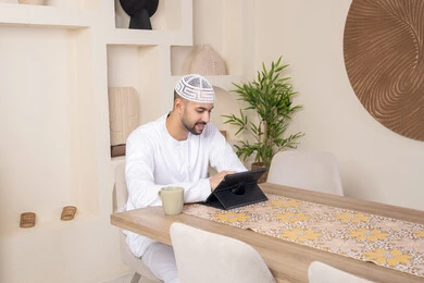 Using a modern, advanced technical device, enjoying hot drinks, performing tasks from home, working remotely, an Emirati Gulf Arab man wearing a kandura and a white taqiya sitting at a table working on a tablet, integrating technology into daily life