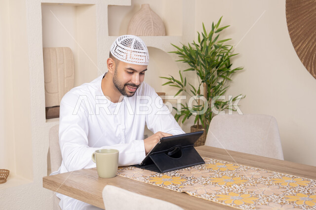 إتمام المهام من المنزل، العمل عن بعد، رجل عربي خليجي اماراتي يرتدي الكندورة والطاقية البيضاء يجلس على الطاولة يعمل على جهاز التابلت، دمج التقنية والتكنولوجيا في الحياة اليومية، استخدام جهاز تقني حديث متطور، الاستمتاع بتناول المشروبات الساخنة