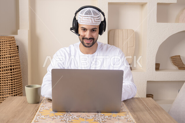 رجل عربي خليجي اماراتي يرتدي الكندورة والهيدفون يعمل على جهاز الحاسوب المحمول في المنزل، الاستماع الى الموسيقى عبر سماعات الاذن، دمج التكنولوجيا والتقنيات الحديثه فى الحياه اليوميه، عقد إجتماع عمل عبر الإنترنت، تناول المشروبات الساخنه