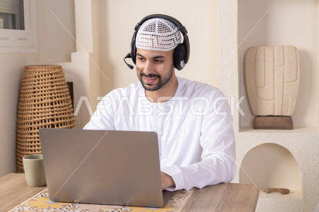 عقد إجتماع عمل عبر الإنترنت، الاستمتاع بالمشروبات الساخنه، رجل عربي خليجي اماراتي يرتدي الكندورة والهيدفون يعمل على جهاز الحاسوب المحمول في المنزل، الاستماع الى الموسيقى عبر سماعات الاذن، دمج التكنولوجيا والتقنيات الحديثه فى الحياه اليوميه