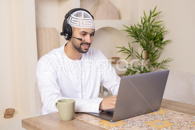 An Emirati Gulf Arab man wearing a kandura and headphones working on his laptop at home, listening to music through earphones, integrating technology and modern techniques into his daily life, holding an online business meeting, drinking hot drinks\