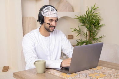 An Emirati Gulf Arab man wearing a kandura and headphones working on his laptop at home, listening to music through earphones, integrating technology and modern techniques into his daily life, holding an online business meeting, drinking hot drinks\