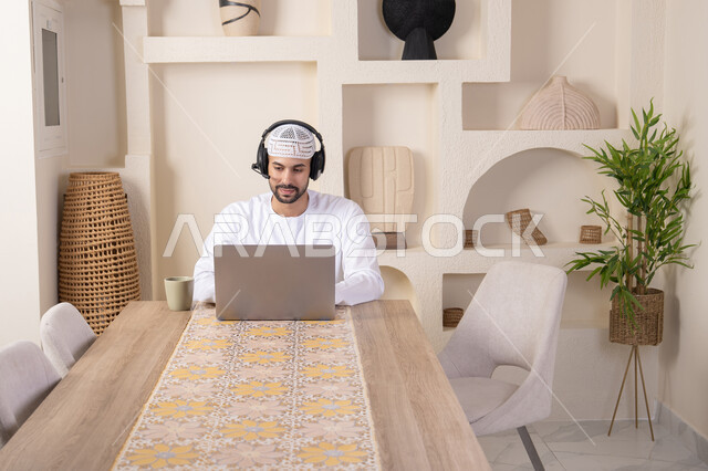 Integrating technology and modern techniques into daily life, holding a business meeting online, enjoying hot drinks, an Emirati Gulf Arab man wearing a kandura and headphones working on his laptop at home, listening to music through earphones