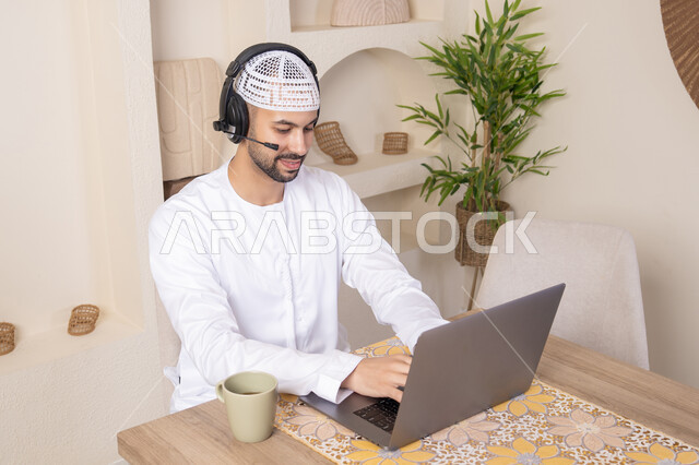 Listening to music through headphones, integrating technology and modern techniques into daily life, holding an online business meeting, enjoying hot drinks, an Emirati Gulf Arab man wearing a kandura and headphones working on his laptop at home