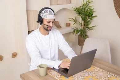 Listening to music through headphones, integrating technology and modern techniques into daily life, holding an online business meeting, enjoying hot drinks, an Emirati Gulf Arab man wearing a kandura and headphones working on his laptop at home
