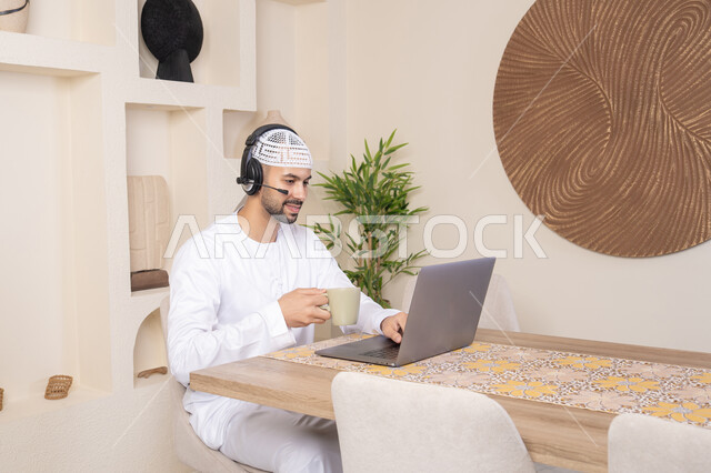 Drinking hot drinks, an Emirati Gulf Arab man wearing a kandura and headphones working on his laptop at home, listening to music through earphones, integrating technology and modern techniques into daily life, holding a business meeting online