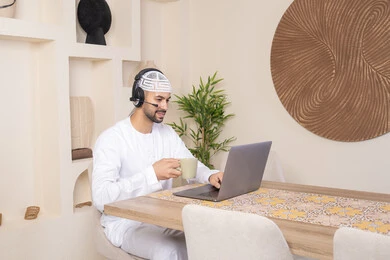 تناول المشروبات الساخنه، رجل عربي خليجي اماراتي يرتدي الكندورة والهيدفون يعمل على جهاز الحاسوب المحمول في المنزل، الاستماع الى الموسيقى عبر سماعات الاذن، دمج التكنولوجيا والتقنيات الحديثه فى الحياه اليوميه، عقد إجتماع عمل عبر الإنترنت