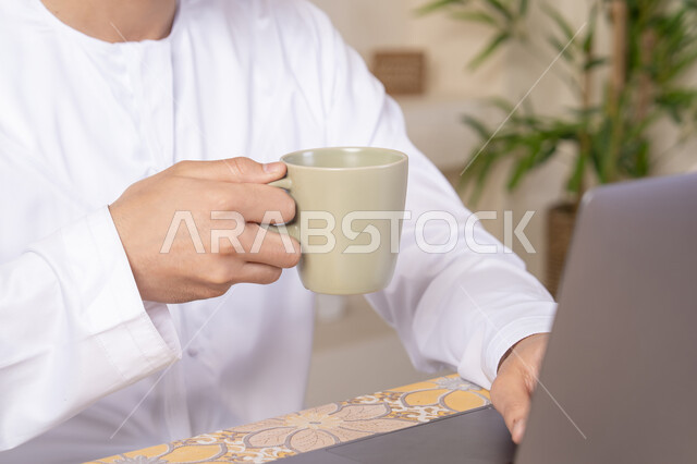 Enjoying hot drinks, an Emirati Gulf Arab man wearing a kandura and headphones working on his laptop at home, listening to music through earphones, integrating technology and modern techniques into daily life, holding a business meeting online