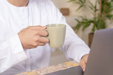 Enjoying hot drinks, an Emirati Gulf Arab man wearing a kandura and headphones working on his laptop at home, listening to music through earphones, integrating technology and modern techniques into daily life, holding a business meeting online