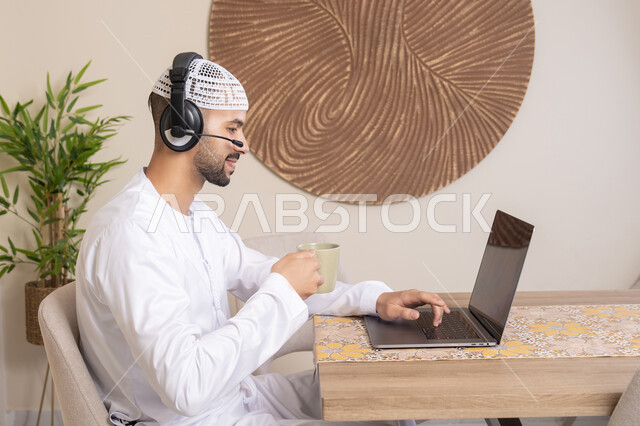 Holding an online business meeting, enjoying hot drinks, an Emirati Gulf Arab man wearing a kandura and headphones working on his laptop at home, listening to music through earphones, integrating technology and modern techniques into daily life
