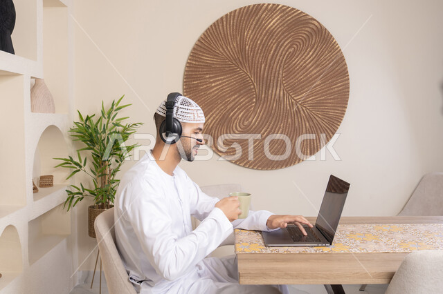 Drinking hot drinks, an Emirati Gulf Arab man wearing a kandura and headphones working on his laptop at home, listening to music through earphones, integrating technology and modern techniques into daily life, holding a business meeting online
