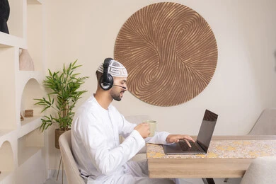 Drinking hot drinks, an Emirati Gulf Arab man wearing a kandura and headphones working on his laptop at home, listening to music through earphones, integrating technology and modern techniques into daily life, holding a business meeting online