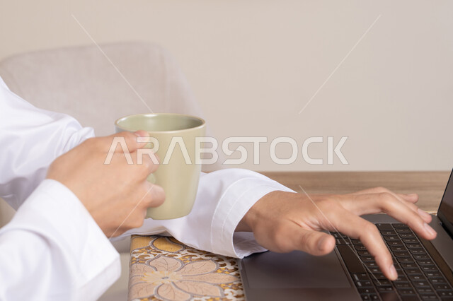 Enjoying hot drinks, an Emirati Gulf Arab man wearing a kandura and headphones working on his laptop at home, listening to music through earphones, integrating technology and modern techniques into daily life, holding a business meeting online