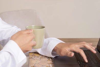 الاستمتاع بالمشروبات الساخنه، رجل عربي خليجي اماراتي يرتدي الكندورة والهيدفون يعمل على جهاز الحاسوب المحمول في المنزل، الاستماع الى الموسيقى عبر سماعات الاذن، دمج التكنولوجيا والتقنيات الحديثه فى الحياه اليوميه، عقد إجتماع عمل عبر الإنترنت