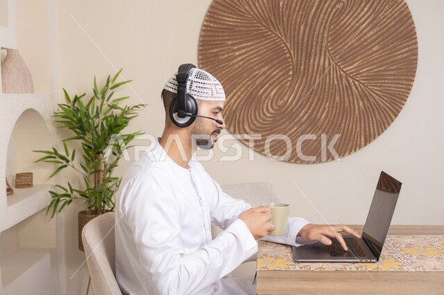 An Emirati Gulf Arab man wearing a kandura and headphones working on his laptop at home, listening to music through earphones, integrating technology and modern techniques into his daily life, holding an online business meeting, drinking hot drinks\