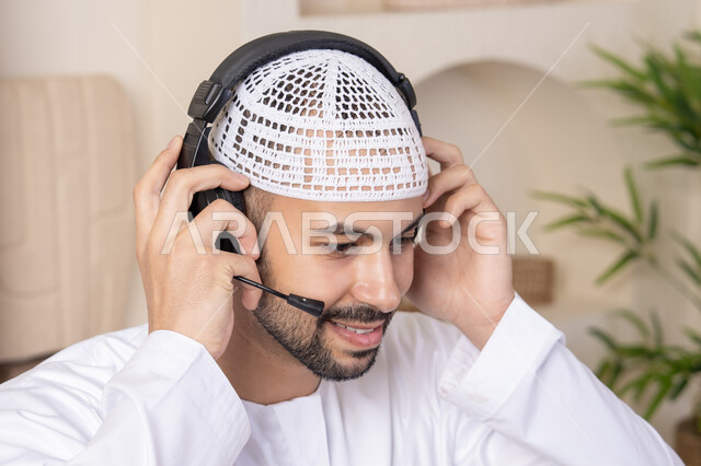 دمج التكنولوجيا والتقنيات الحديثه فى الحياه اليوميه، عقد إجتماع عمل عبر الإنترنت، الاستمتاع بالمشروبات الساخنه، رجل عربي خليجي اماراتي يرتدي الكندورة والهيدفون يعمل على جهاز الحاسوب المحمول في المنزل، الاستماع الى الموسيقى عبر سماعات الاذن