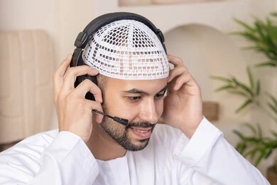 Integrating technology and modern techniques into daily life, holding a business meeting online, enjoying hot drinks, an Emirati Gulf Arab man wearing a kandura and headphones working on his laptop at home, listening to music through earphones