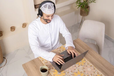 Listening to music through headphones, integrating technology and modern techniques into daily life, holding an online business meeting, enjoying hot drinks, an Emirati Gulf Arab man wearing a kandura and headphones working on his laptop at home