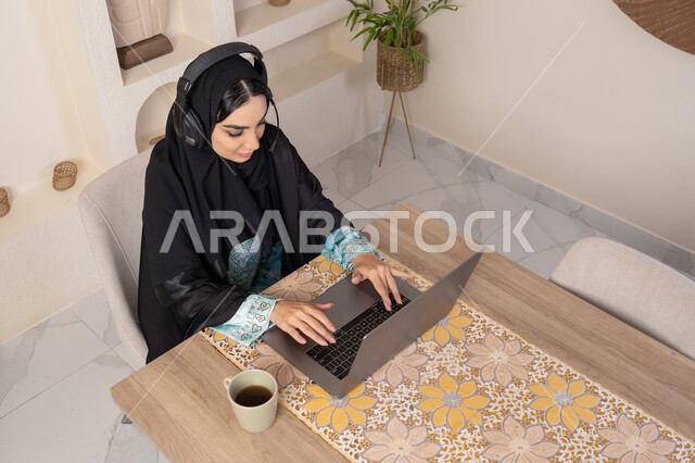 دمج التكنولوجيا الحديثة مع الروتين اليومي، نمط حياة صحي، امراه عربية خليجية سعودية ترتدي العباءة والحجاب تجلس على طاولة في المنزل تستخدم الحاسب المحمول، سيدة اماراتية تضع الهيدفون ، أجواء صباحية هادئة في المنزل