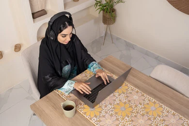 Integrating modern technology with daily routine, healthy lifestyle, a Saudi Arabian Gulf woman wearing an abaya and hijab sitting at a table at home using a laptop, an Emirati woman wearing headphones, a calm morning atmosphere at home