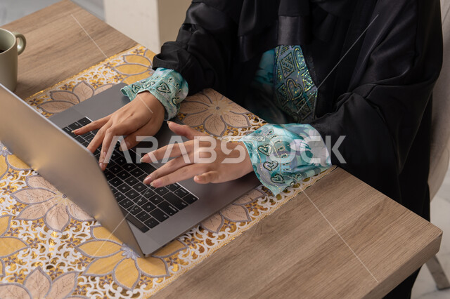 الاستمتاع بشرب القهوة، دمج التكنولوجيا الحديثة مع الروتين اليومي، نمط حياة صحي، امراه عربية خليجية سعودية ترتدي العباءة والحجاب تجلس على طاولة في المنزل تستخدم الحاسب المحمول، سيدة اماراتية تضع الهيدفون ، أجواء صباحية هادئة في المنزل